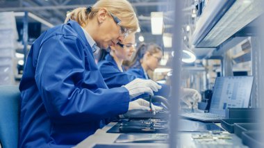 Mavi İş Ceketi ve Koruyucu Gözlüklü Kadın Elektronik Fabrikası İşçisi, Akıllı Telefonları Tornavidayla Birleştiriyor. Arka planda daha fazla çalışanı olan Yüksek Teknoloji Fabrikası.