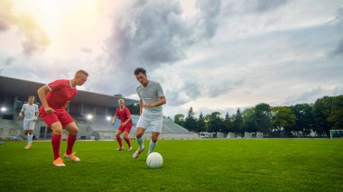 Profesyonel Futbolcu Topla Öncülük Ediyor, Rakiplerinin Kayan Aletleri Ustaca Sürüyor ve Baypas Ediyor. İki Profesyonel Futbol Takımı Oynuyor. Düşük açılı atış. Sıcak Güneş Işığı Parıltısı.