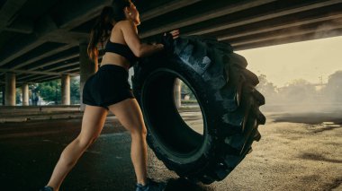Siyah Atletizm Üstündeki Güzel Kendine Güvenen Fitness Kızı Sokakta Egzersiz Yapıyor. Kentsel bir ortamda Büyük Ağır Bir Tekerleği Döndürüyor.