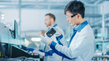 Yüksek Teknoloji Fabrikası: Kalite Kontrol Mühendisi Akıllı Elektronik Cihazı alıp olası hasarlar için görsel olarak kontrol ediyor. PCB için Yüzey Mount Pick ve Yerleştirme Arkaplan Assembly Line