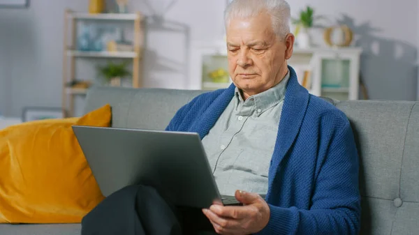 Hombre mayor progresivo en casa sentado en el sofá utiliza el ordenador portátil. Lleno de vida Anciano Relajándose en casa, leyendo noticias en el cuaderno. — Foto de Stock