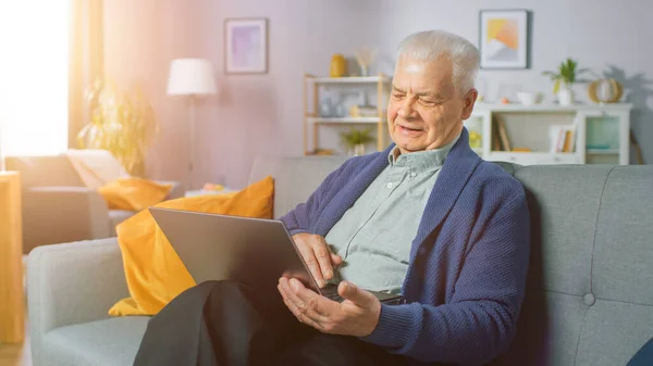 Hombre mayor progresivo en casa sentado en el sofá utiliza el ordenador portátil. Lleno de vida Anciano Relajándose en casa, leyendo noticias en Internet. — Foto de Stock