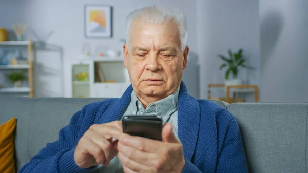 Hombre mayor progresivo sentado en su sala de estar utiliza fácilmente el teléfono inteligente, hace gestos conmovedores y se siente muy cómodo con las nuevas tecnologías. — Foto de Stock