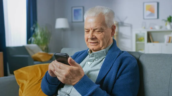 Retrato del hombre mayor progresivo sentado en su sala de estar utiliza fácilmente el teléfono inteligente, hace gestos conmovedores y se siente muy cómodo con las nuevas tecnologías. — Foto de Stock