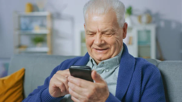 Retrato del hombre mayor progresivo sentado en su sala de estar utiliza fácilmente el teléfono inteligente, hace gestos conmovedores y se siente muy cómodo con las nuevas tecnologías. — Foto de Stock
