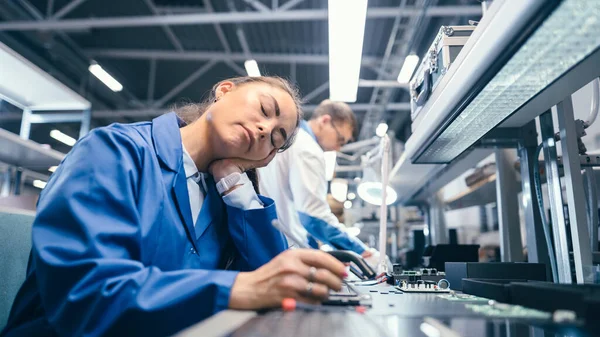 Fotografía de una mujer cansada durmiendo vestida con un abrigo de trabajo azul en su lugar de trabajo en la fábrica de electrónica. Fábrica de alta tecnología con más empleados en segundo plano. —  Fotos de Stock