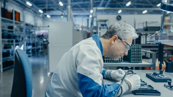 Senior Man in Blue - White Work Coat używa agregatu Plier do montażu płytek obwodów drukowanych dla smartfona. Robotnicy fabryki elektroniki w obiekcie High Tech Factory. — Zdjęcie stockowe