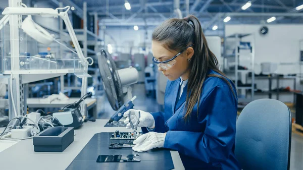 Young Female Blue and White Work Coat está utilizando Plier para montar la placa de circuito impreso para Smartphone. Trabajadores de la fábrica de electrónica en una instalación de fábrica de alta tecnología. — Foto de Stock