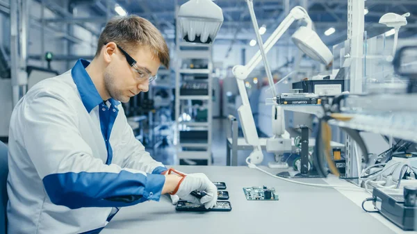 Młody człowiek w niebiesko-białym płaszczu roboczym używa Plier do montażu płytek obwodów drukowanych dla smartfona. Robotnicy fabryki elektroniki w obiekcie High Tech Factory. — Zdjęcie stockowe