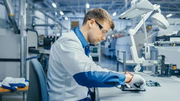 El joven con abrigo de trabajo azul y blanco está usando Plier para ensamblar la placa de circuito impreso para Smartphone. Trabajadores de la fábrica de electrónica en una instalación de fábrica de alta tecnología. — Foto de Stock