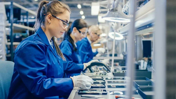 2015 년 12 월 1 일에 확인 함 . Female Electronics Factory Workers in Blue Work Coat and Protective Glasses Assembling Printed Circuit Boards for Smartphone with Tweezers. 배경에 더 많은 직원이 있는 첨단 기술 공장. — 스톡 사진