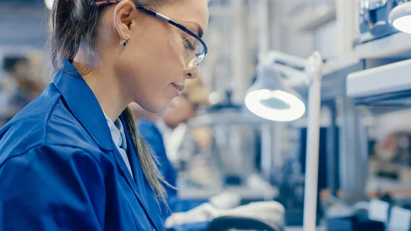 ブルーワークコートと保護メガネの女性電子工場労働者はピンセットとドライバーでスマートフォンを組み立てています。背景により多くの従業員を抱えるハイテク工場. — ストック写真