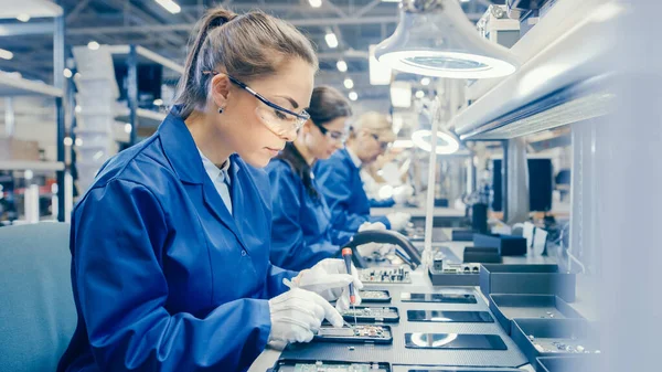 Жінка-фабрика електроніки Працівниця в синьому одязі та захисних окулярах збирає смартфони разом з Screwdriver. Висока технічна фабрика з більшою кількістю робітників на задньому плані. — стокове фото