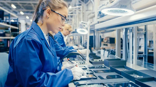 Γυναίκα Εργάτης Εργοστάσιο Ηλεκτρονικών σε μπλε παλτό εργασίας και προστατευτικά γυαλιά είναι Συνέλευση Smartphones με κατσαβίδι. Εγκαταστάσεις υψηλής τεχνολογίας εργοστάσιο με περισσότερους εργαζόμενους στο παρασκήνιο. — Φωτογραφία Αρχείου