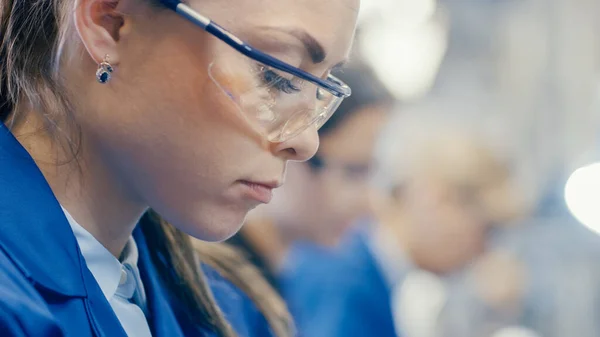 Närbild Skott av en kvinnlig Electronics Factory Worker i blått arbete Coat och skyddsglasögon tittar ner och arbetar. Högteknologisk fabrik med flera anställda. — Stockfoto