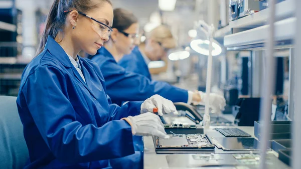 Female Electronics Factory Worker in Blue Work Coat and Protective Glasses is assembling Laptops Motherboard with a Screwdriver. Zařízení High Tech Factory s více zaměstnanci. — Stock fotografie