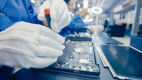 Közelkép egy női elektronikai gyári munkásról kék munkásköpenyben Laptopok összeszerelése Alaplap csavarhúzóval. High Tech Factory létesítmény több alkalmazottal. — Stock Fotó