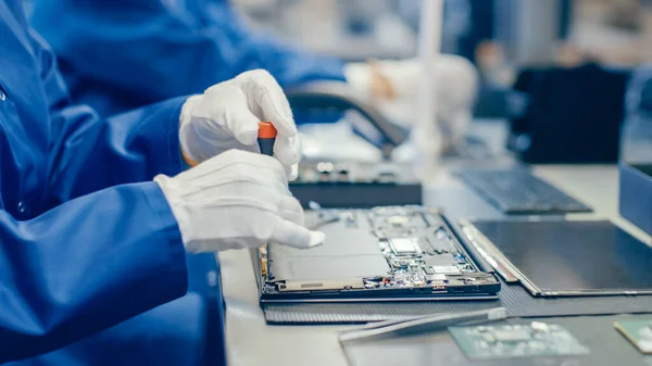 Close-up de uma funcionária feminina de fábrica de eletrônicos em azul casaco de trabalho montagem de laptops placa-mãe com uma chave de fenda. Instalação de fábrica de alta tecnologia com vários funcionários. — Fotografia de Stock