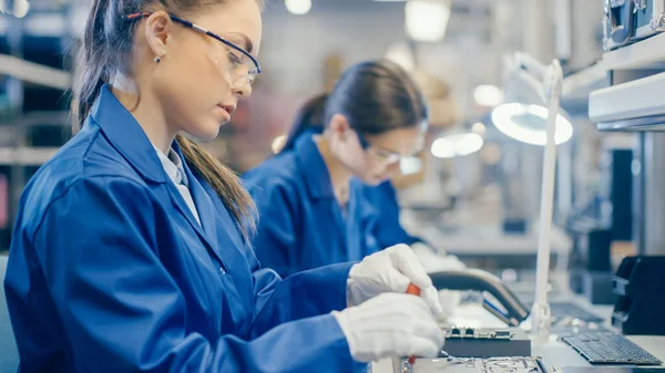 Eine Arbeiterin in einer Elektronikfabrik in blauem Arbeitsmantel und Schutzbrille montiert mit einem Schraubenzieher ein Laptop-Mainboard. High-Tech-Fabrik mit mehreren Mitarbeitern. — Stockfoto