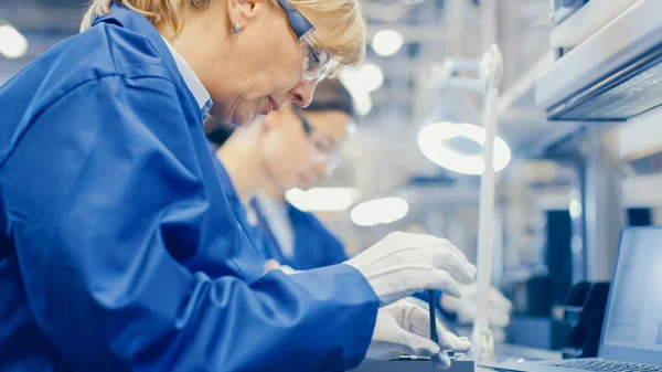 Eine Arbeiterin in einer Elektronikfabrik in blauem Arbeitsmantel und Schutzbrille montiert Smartphones mit Schraubenzieher. High-Tech-Fabrik mit mehr Mitarbeitern im Hintergrund. — Stockfoto