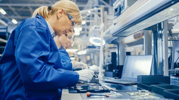 La trabajadora femenina de la fábrica de electrónica con capa de trabajo azul y gafas protectoras está ensamblando teléfonos inteligentes con destornillador. Fábrica de alta tecnología con más empleados en segundo plano. — Foto de Stock