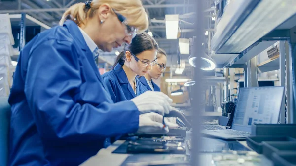Kvinnliga Electronics Factory Worker i blått arbete Coat och skyddsglasögon är att montera Smartphones med skruvmejsel. High Tech Factory anläggning med fler anställda i bakgrunden. — Stockfoto