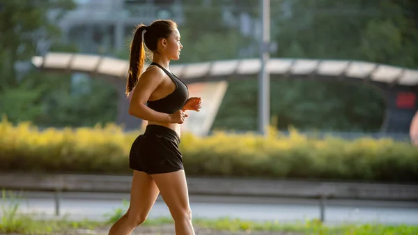 Mooie rondborstige fitness meisje in zwart atletische Top en Shorts is energiek Running in de straat. Ze is aan het joggen in een stedelijke omgeving onder een brug. — Stockfoto