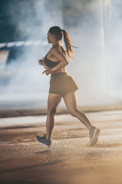 Backshot di una ragazza forte fitness in Top atletico nero e pantaloncini da jogging su una strada. L'atleta corre in un ambiente urbano sotto un ponte. — Foto Stock