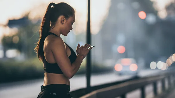 Vista laterale Colpo di una bella ragazza di fitness sicura in Top atletico nero sta utilizzando uno smartphone su una strada. Lei è in un ambiente urbano sotto un ponte. — Foto Stock