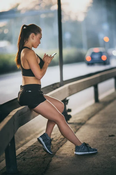 Shot of a Beautiful Confident Fitness Girl in Black Athletic Top utilise un smartphone dans une rue. Elle est dans un environnement urbain sous un pont. — Photo