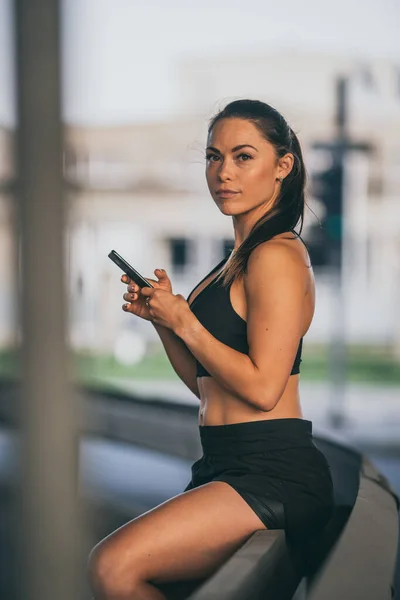 Beautiful Confident Fitness Girl in Black Athletic Top utilise un smartphone dans une rue et regarde la caméra. Elle est dans un environnement urbain sous un pont avec des voitures en arrière-plan. — Photo