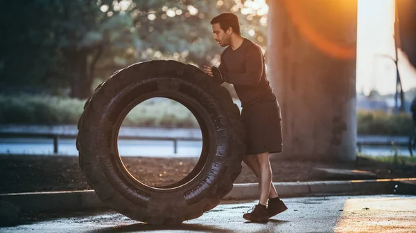 Forte Muscular Fit Young Man está fazendo exercícios na rua. Levantando e rolando um pneu pesado grande em um ambiente urbano. — Fotografia de Stock