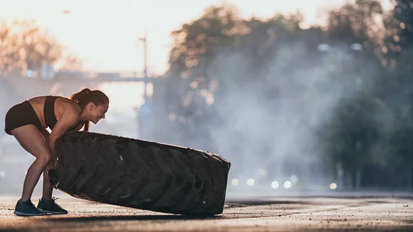 Девушка в черном спортивном топе и шортах делает упражнения на улице. Прокат большой тяжелой шины в городской среде — стоковое фото