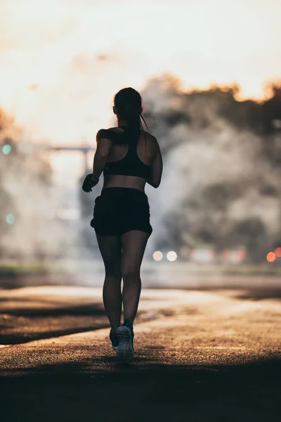 Backshot di una ragazza forte fitness in Top atletico nero e pantaloncini da jogging su una strada. L'atleta sta correndo in un ambiente urbano sotto un ponte con ambiente oscuro. — Foto Stock