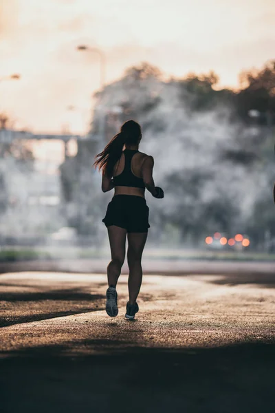 Backshot of a Strong Fitness Girl in Black Athletic Top and Shorts Jogging on a Dark Foggy Street. L'athlète court dans un environnement urbain sous un pont. — Photo