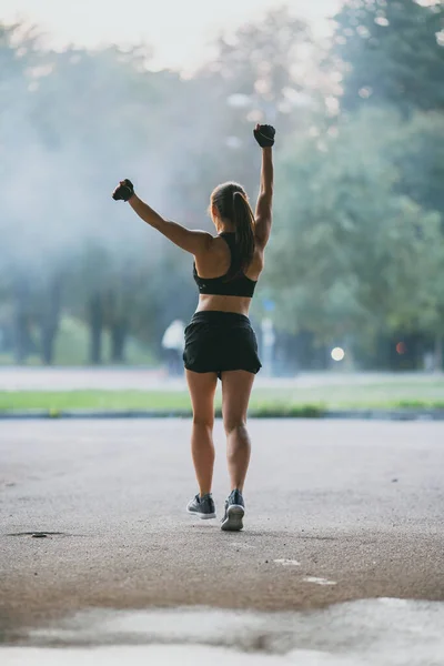 Backshot di una ragazza forte fitness in Top atletico nero e pantaloncini da jogging al mattino su una Foggy Street. Atleta sta correndo in un ambiente urbano e allungando le mani. — Foto Stock