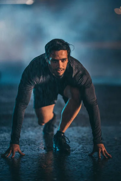 Forte Muscular Fit Young Man Pronto para Sprint em uma noite chuvosa. Ele está treinando em um ambiente urbano sob uma ponte com fundo nebuloso escuro. — Fotografia de Stock