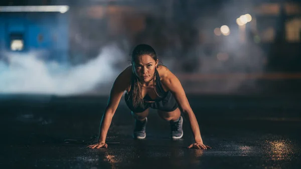 Beautiful Energetic Fitness Girl en camiseta y pantalones cortos deportivos negros está haciendo ejercicios push up. Ella está haciendo un entrenamiento en un ambiente urbano húmedo y nebuloso debajo de un puente en el fondo. — Foto de Stock