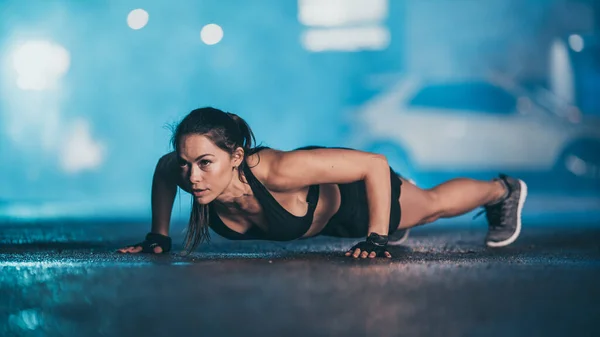 Gyönyörű Energetikai Fitness Girl fekete atlétikai Top és rövidnadrág csinál push up gyakorlatok. Edzést tart egy esténként nedves városi környezetben köddel és autókkal a háttérben.. — Stock Fotó