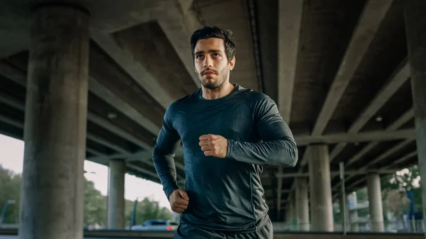 Athletic Young Man in Sports Outfit is Jogging in the Street. Ele está correndo em um ambiente urbano sob uma ponte com carros no fundo. — Fotografia de Stock