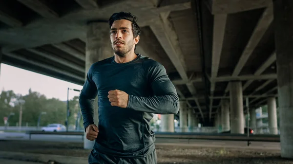 Athletic Young Man in Sports Outfit is Jogging in the Street. Ele está correndo em um ambiente urbano sob uma ponte com carros no fundo. — Fotografia de Stock