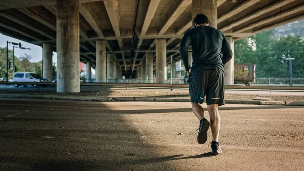 Backshot de um jovem atlético em trajes esportivos Jogging in the Street. Ele está correndo em um ambiente urbano sob uma ponte com carros no fundo. — Fotografia de Stock