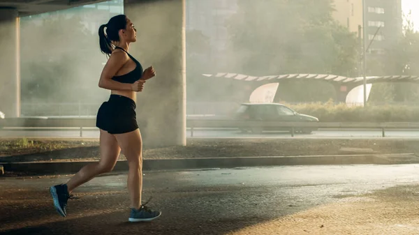 Girato di una bella ragazza di forma fisica in Top atletico nero e pantaloncini da jogging in strada con luce calda. Sta correndo in un ambiente urbano sotto un ponte con le auto sullo sfondo. — Foto Stock