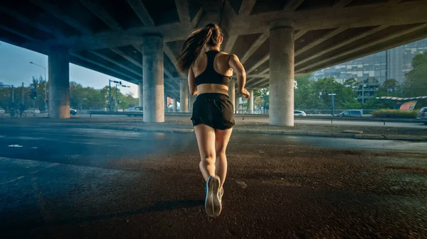 Backshot av en stark Fitness Girl i svart atletisk topp och shorts jogging genom en rökig gata. Hon springer i en stadsmiljö under en bro med bilar i bakgrunden. — Stockfoto