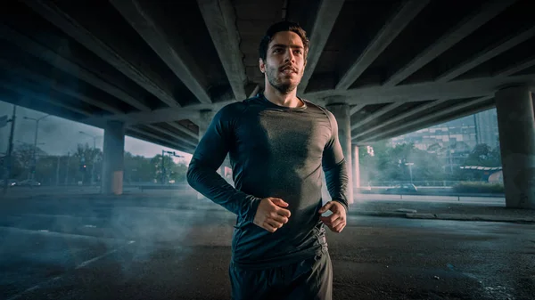 거리에서 조깅하는 운동 선수의 근육질의 젊은 남자의 초상화 사진. 그는 다리 밑의 도시 환경에서 달리고 있다. — 스톡 사진
