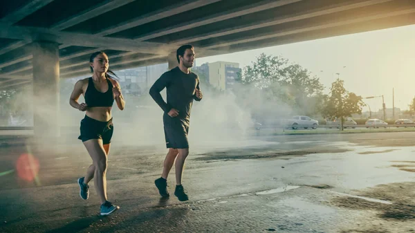 Starka och vackra Athletic Fitness par i träningskläder Jogga på en gata fylld med Steam på en solig dag. De springer i en stadsmiljö under en bro med bilar i — Stockfoto