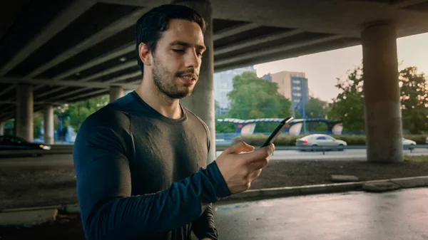 Sportlich muskulöser junger Mann in Sportkleidung joggt in einer Straße. Er benutzt sein Smartphone. Sportler läuft in urbaner Umgebung unter einer Brücke mit Autos im Hintergrund. — Stockfoto