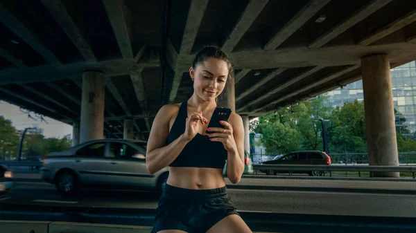 Bela garota de fitness confiante em Black Athletic Top está usando um smartphone em uma rua. Ela está em um ambiente urbano sob uma ponte com carros no fundo. — Fotografia de Stock