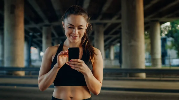 Belle fille de remise en forme forte en noir Athletic Top utilise un smartphone et souriant sur une rue. Elle est dans un environnement urbain sous un pont. — Photo