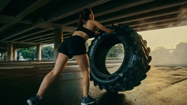 Schöne selbstbewusste Fitness-Mädchen in schwarzem athletischen Top macht Übungen auf der Straße. Schuppen kippen einen großen schweren Reifen in einer städtischen Umgebung. — Stockfoto
